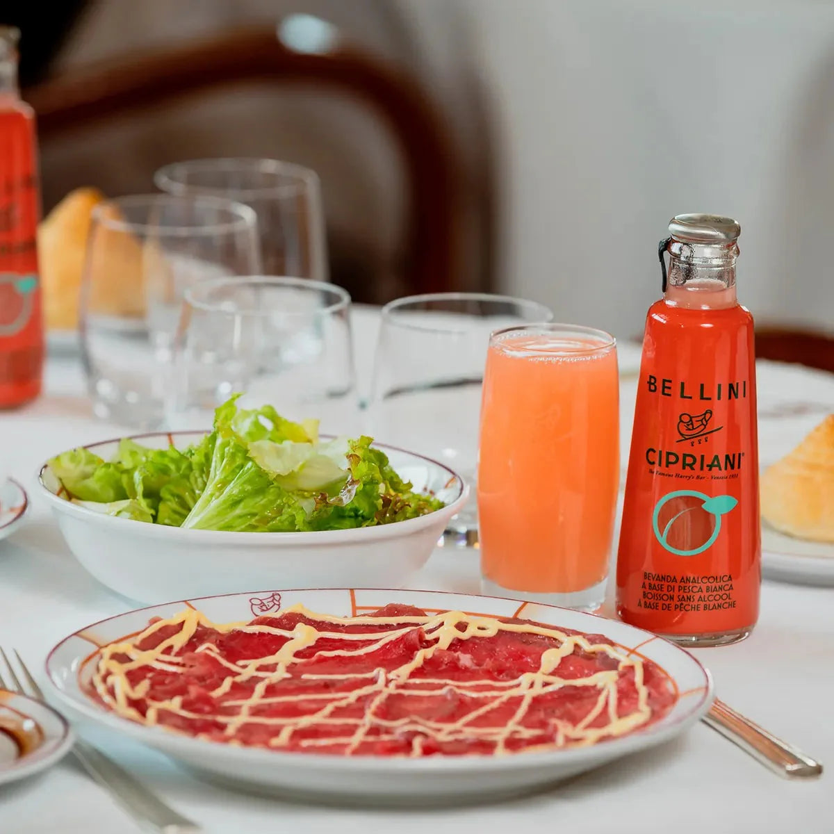 Lifestyle photo of Cipriani White Peach Virgin Bellini with glass, salad, and carpaccio
