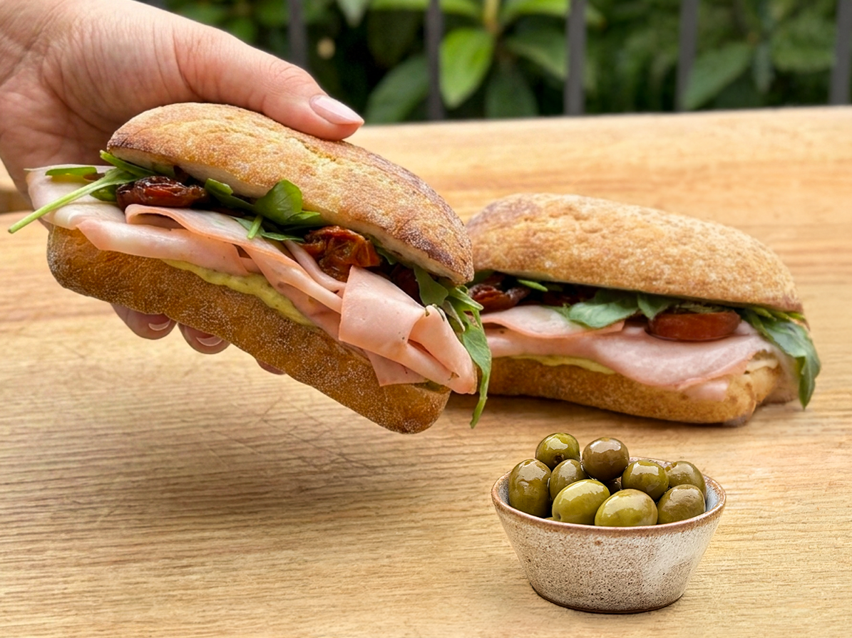 Gourmet Italian sandwich ingredients including sliced mortadella, fresh mozzarella, Roi basil pesto, and Cutrera sun-dried tomatoes.