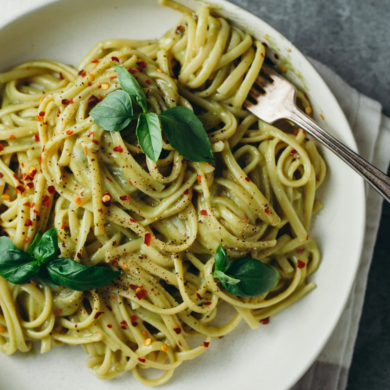 A close-up overhead shot of a white plate filled with creamy green pasta, garnished with fresh basil leaves and red chili flakes, with a fork resting on the side, set on a gray textured surface with a folded white napkin.