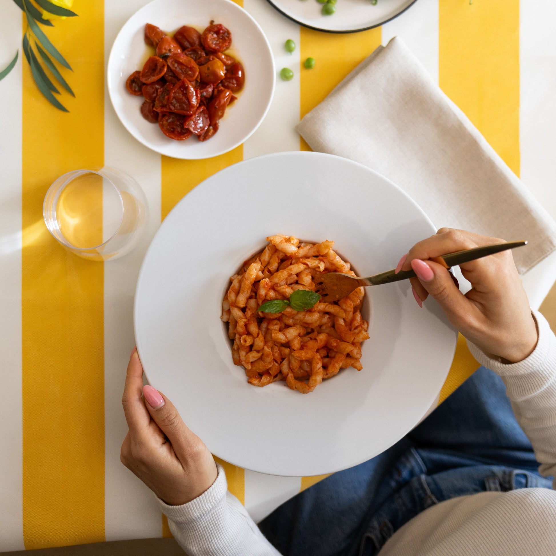 Overhead view of a person holding a white plate of pasta with tomato sauce, with a fork in their hand, set on a yellow and white striped tablecloth with a glass of water and a side dish of sunflower seeds.