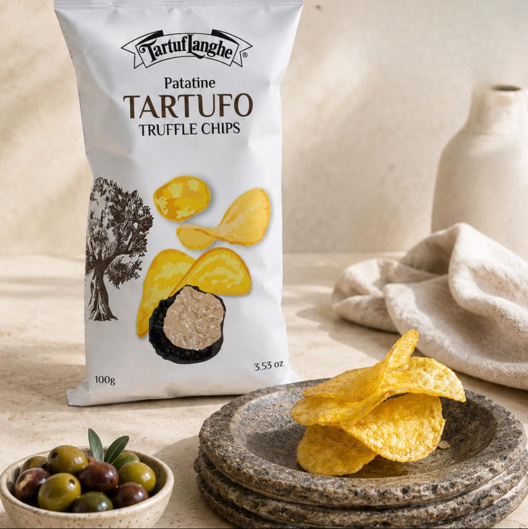 A bag of Tartufange Patatine Tartufo Truffle Chips stands on a stone surface with loose chips in a dish, olives in a small bowl, and a neutral ceramic vase in the background. Text at the top reads 'Elevated Snacking Experience' and 'CRUMBLE OVER SALADS OR PAIR WITH SPREADS.