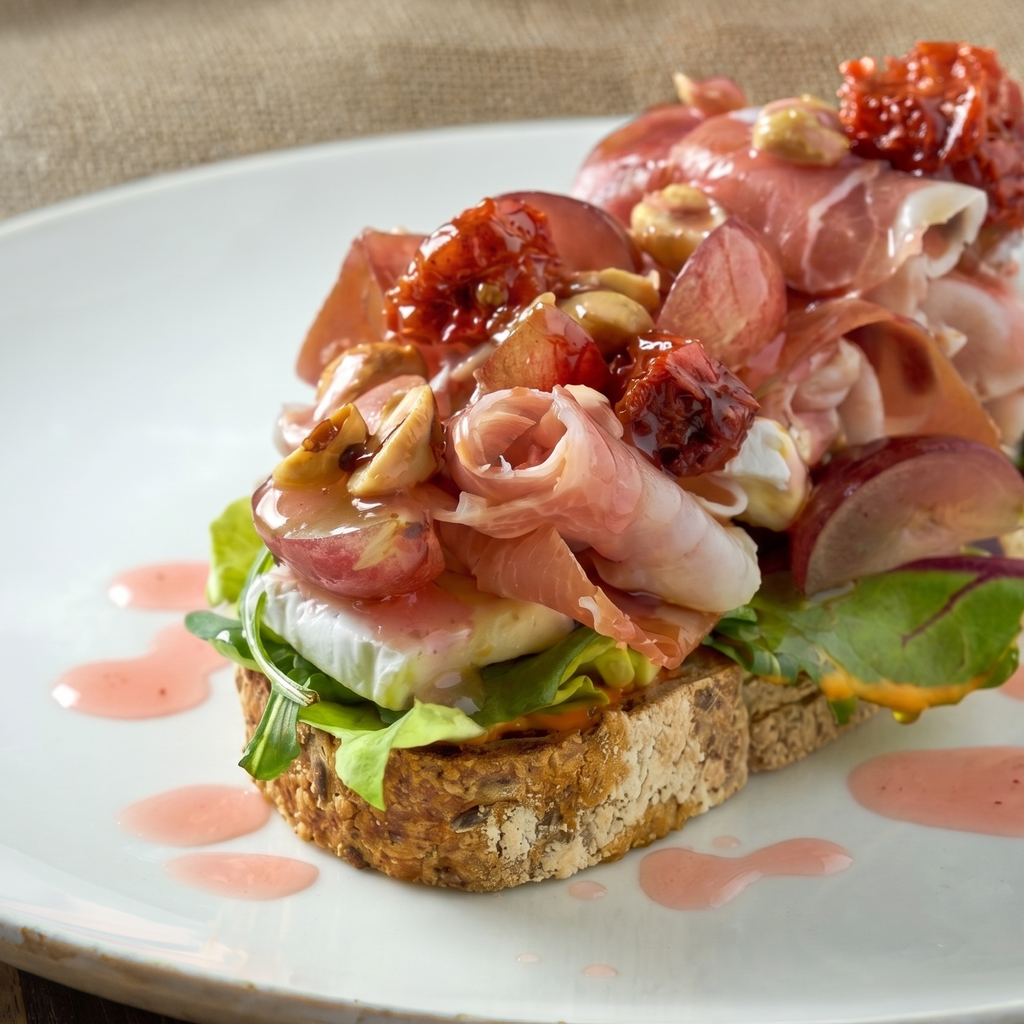 Two slices of toasted bread topped with prosciutto crudo, white cheese, red onion, nuts, and dried fruit, served on a white plate with pink dressing drizzle.