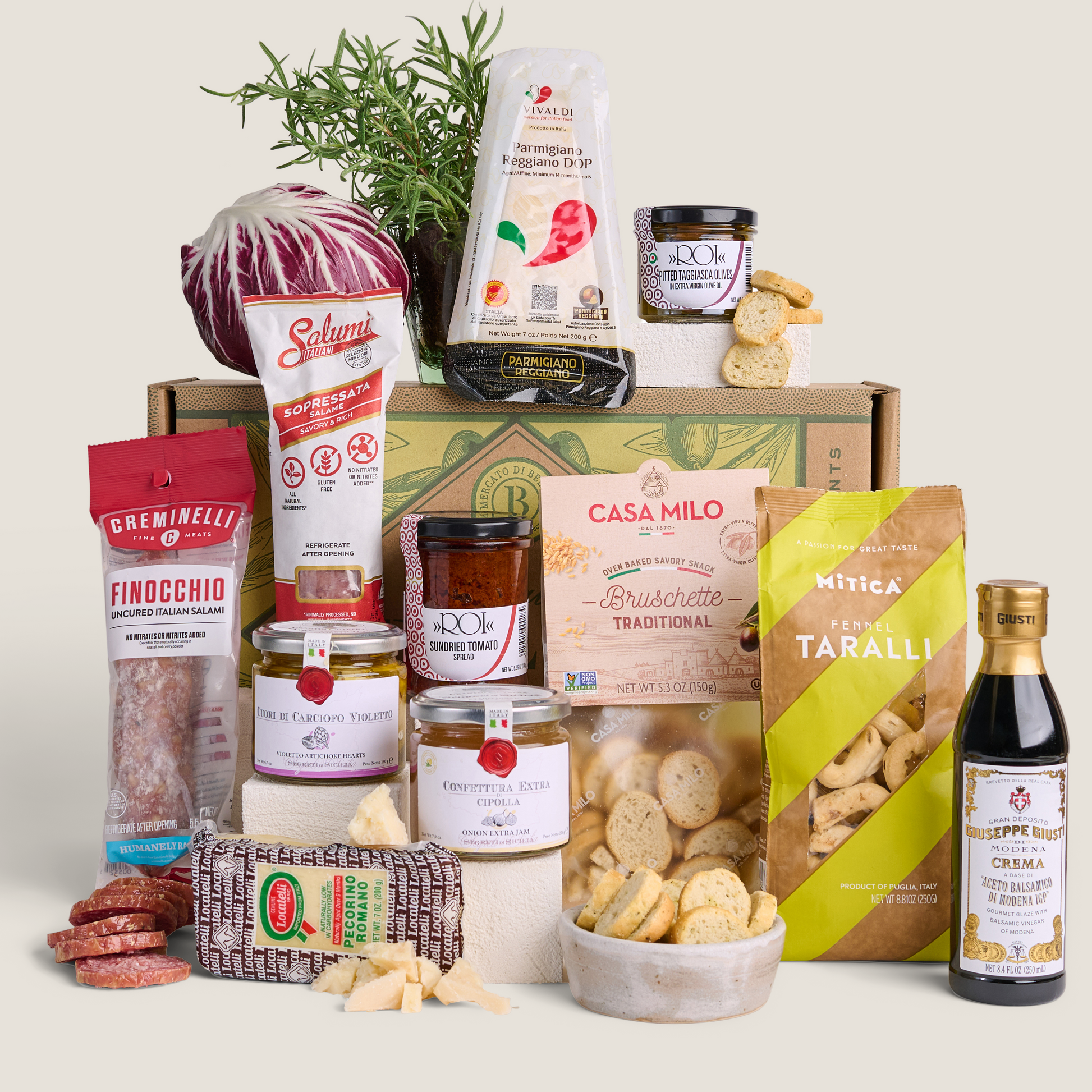 A curated gourmet food gift box featuring an assortment of Italian products including cured meats, cheeses, crackers, and a bottle of balsamic vinegar, arranged in front of a branded cardboard box on a solid light beige background.