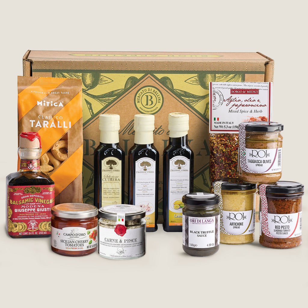 A curated gourmet gift box set featuring olive oil, balsamic vinegar, spreads, and snacks arranged in front of a branded gift box on a light beige background.