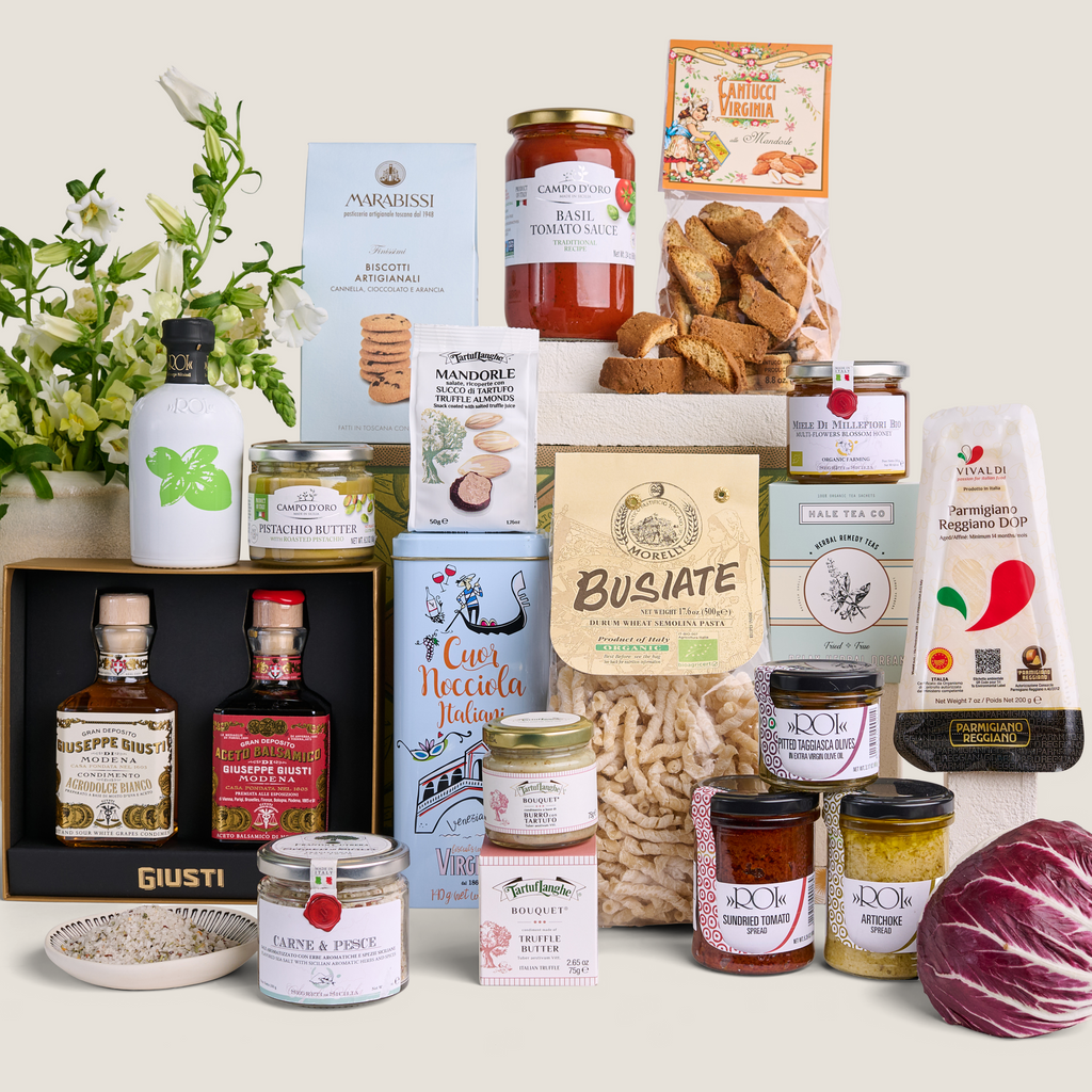 A curated gourmet food and beverage gift assortment arranged on a light beige background. The collection includes artisanal cheeses, cured meats, pasta, preserves, crackers, tea, and spirits in branded packaging, with a small white flower arrangement on the left and a red cabbage on the bottom right.