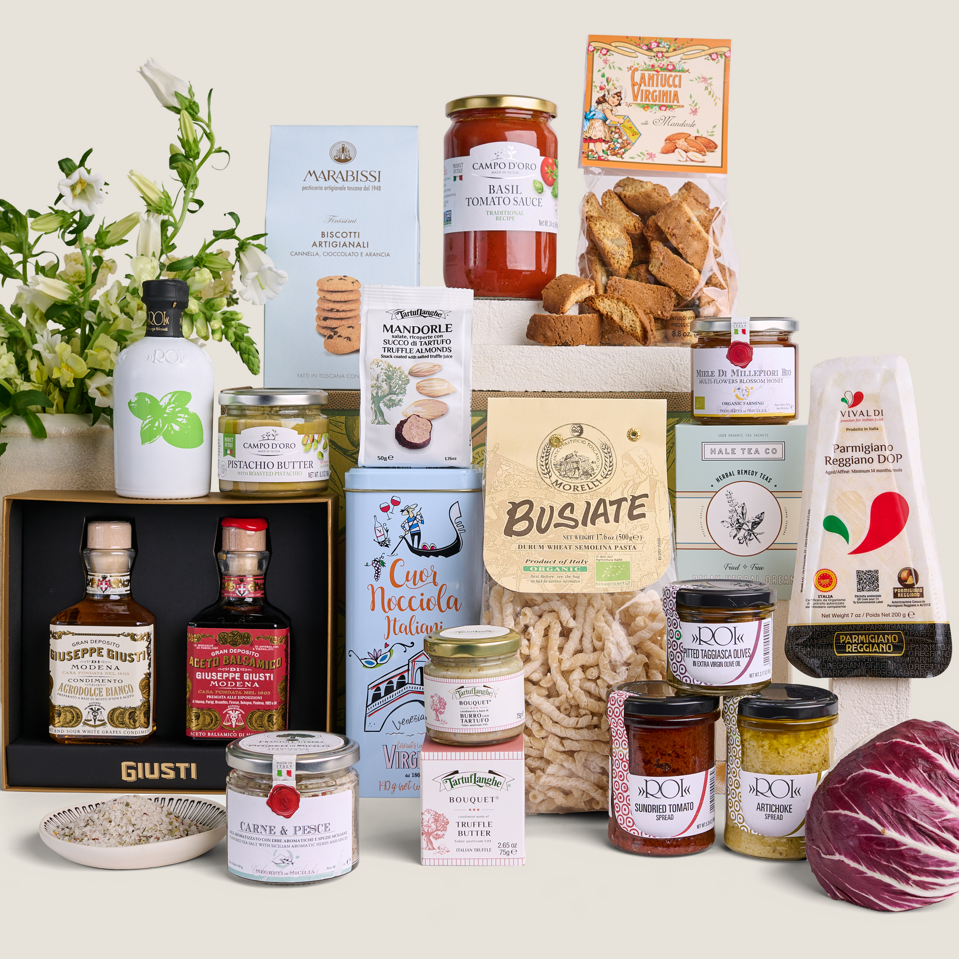 A curated gourmet food and beverage gift assortment arranged on a light beige background. The collection includes artisanal cheeses, cured meats, pasta, preserves, crackers, tea, and spirits in branded packaging, with a small white flower arrangement on the left and a red cabbage on the bottom right.