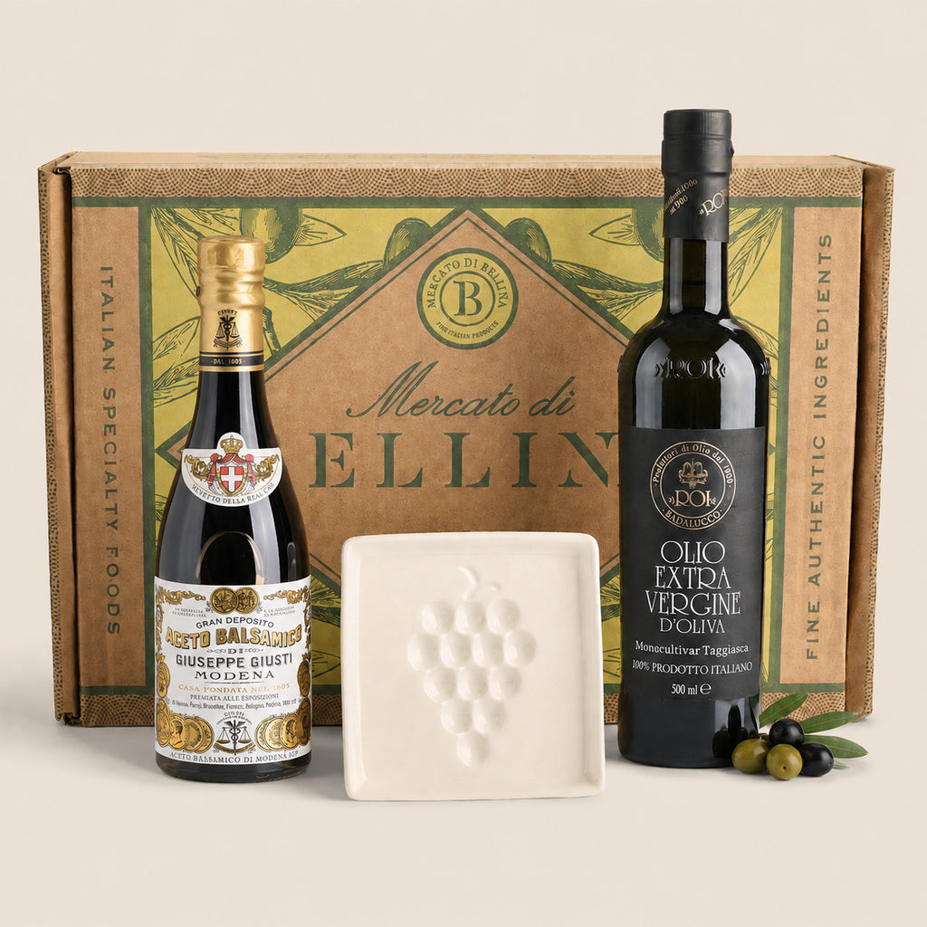 A gift set featuring a bottle of Edo Balsamino Siciliano grapefruit soda, a bottle of Mercato di Ellina extra virgin olive oil, a white square ceramic plate with a grape motif, and a green and brown gift box, arranged on a light beige background.