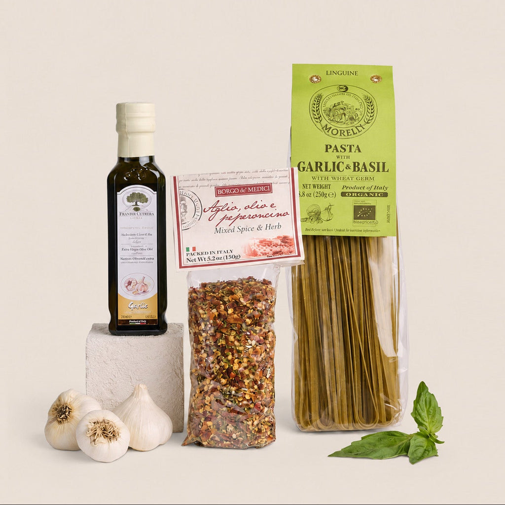 A styled food product set on a warm off-white background, featuring a bottle of olive oil on a small pedestal, a bag of spaghetti, a bag of garlic and basil seasoning mix, whole garlic bulbs, and fresh basil leaves.