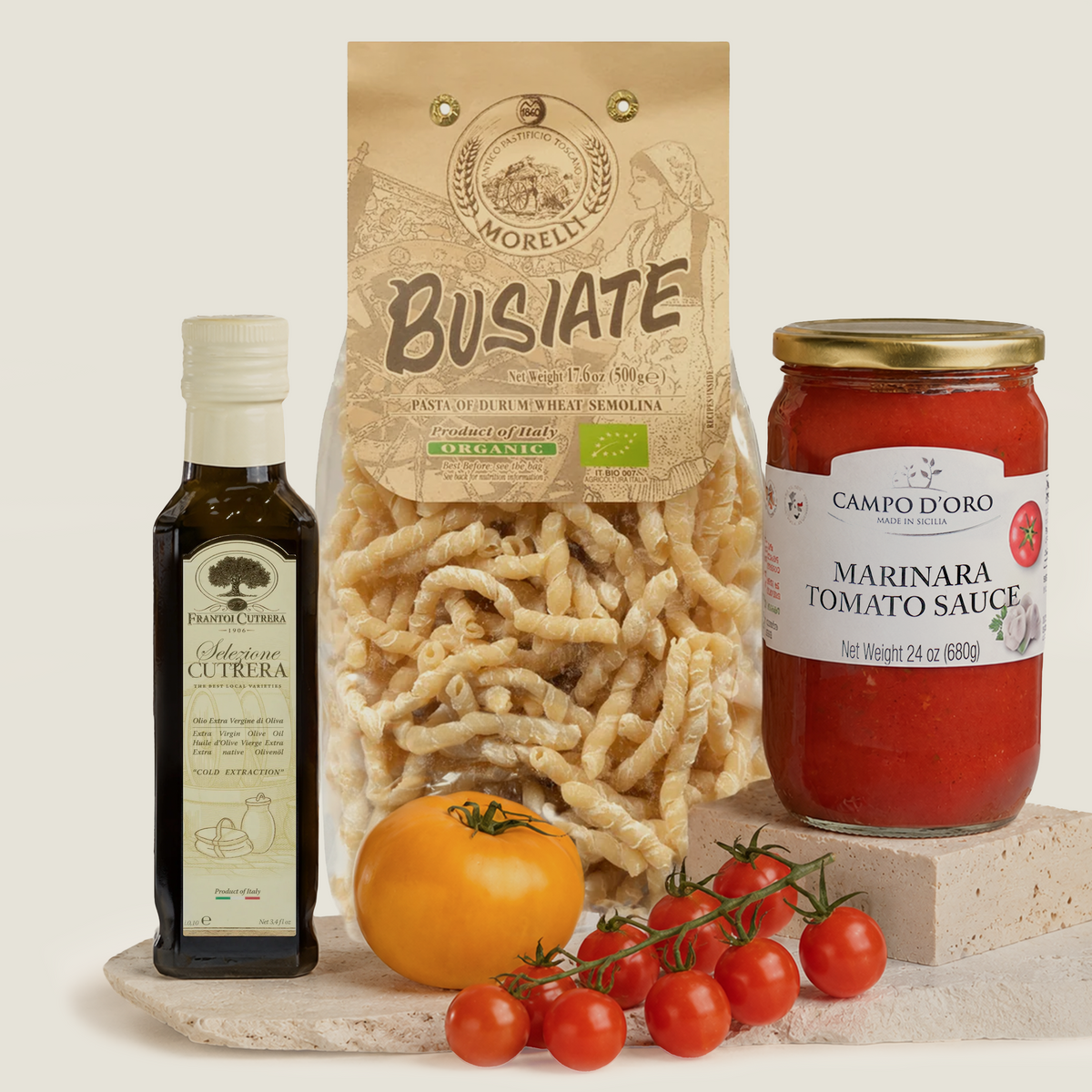A styled product shot featuring a bag of Morelli Busiate pasta, a bottle of Frantoi Cutrera Selezione olive oil, a jar of Campo d'Oro Marinara Tomato Sauce, a whole tomato, and a cluster of cherry tomatoes on a stone slab against a light beige background.