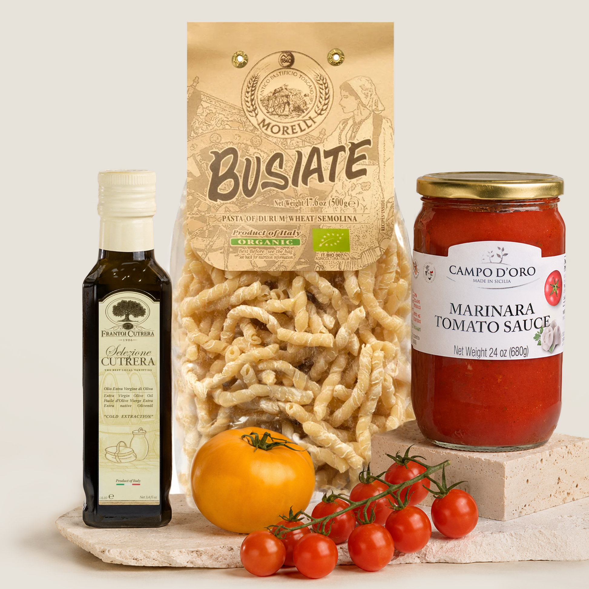 A styled product shot featuring a bag of Morelli Busiate pasta, a bottle of Frantoi Cutrera Selezione olive oil, a jar of Campo d'Oro Marinara Tomato Sauce, a whole tomato, and a cluster of cherry tomatoes on a stone slab against a light beige background.