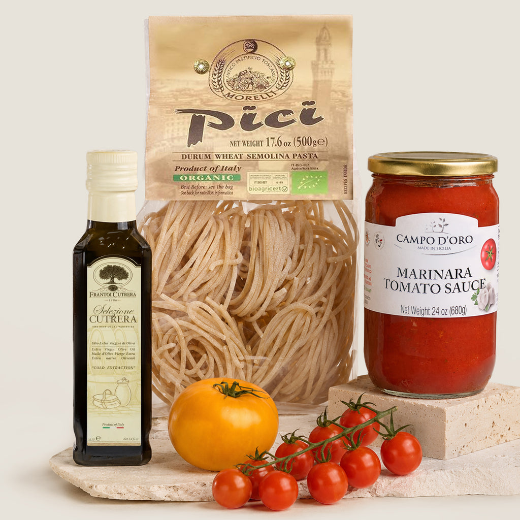 A styled product shot featuring a bag of Morelli Busiate pasta, a bottle of Frantoi Cutrera Selezione olive oil, a jar of Campo d'Oro Marinara Tomato Sauce, a whole tomato, and a cluster of cherry tomatoes on a stone slab against a light beige background.