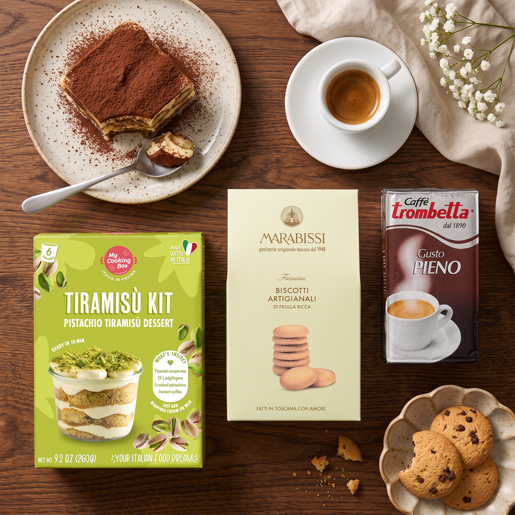 A styled flat-lay of a tiramisu recipe kit on a wooden table, featuring a green Marabissi Tiramisñu Kit box, a cream-colored Marabissi Biscotti Artigianali box, and a Gaffé Trombetta Caffé Pièno coffee box, surrounded by food props like a slice of tiramisu, a cup of espresso, cookies, and a cloth.