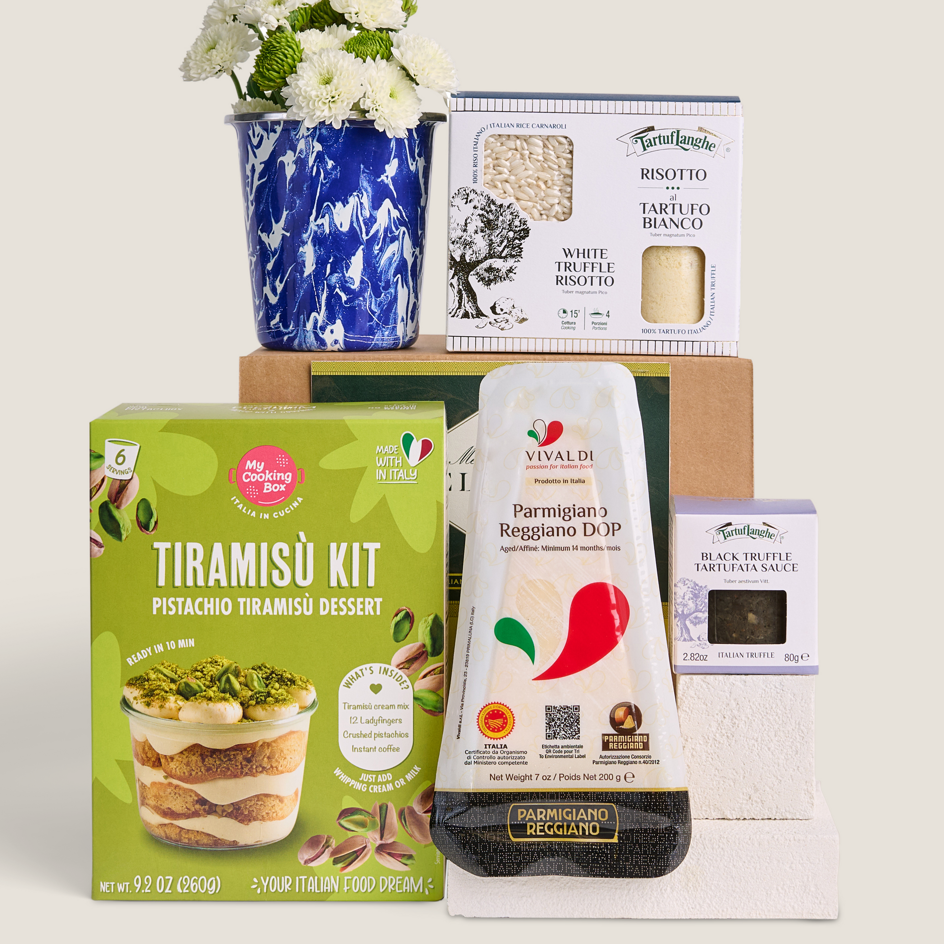 A curated gift set featuring a green Tiramisu Kit box, a white and red Parmigiano cheese block, a white truffle risotto kit, black tartufata sauce, a blue-and-white floral ceramic vase with white flowers, and a brown cardboard box, arranged on a light beige background.