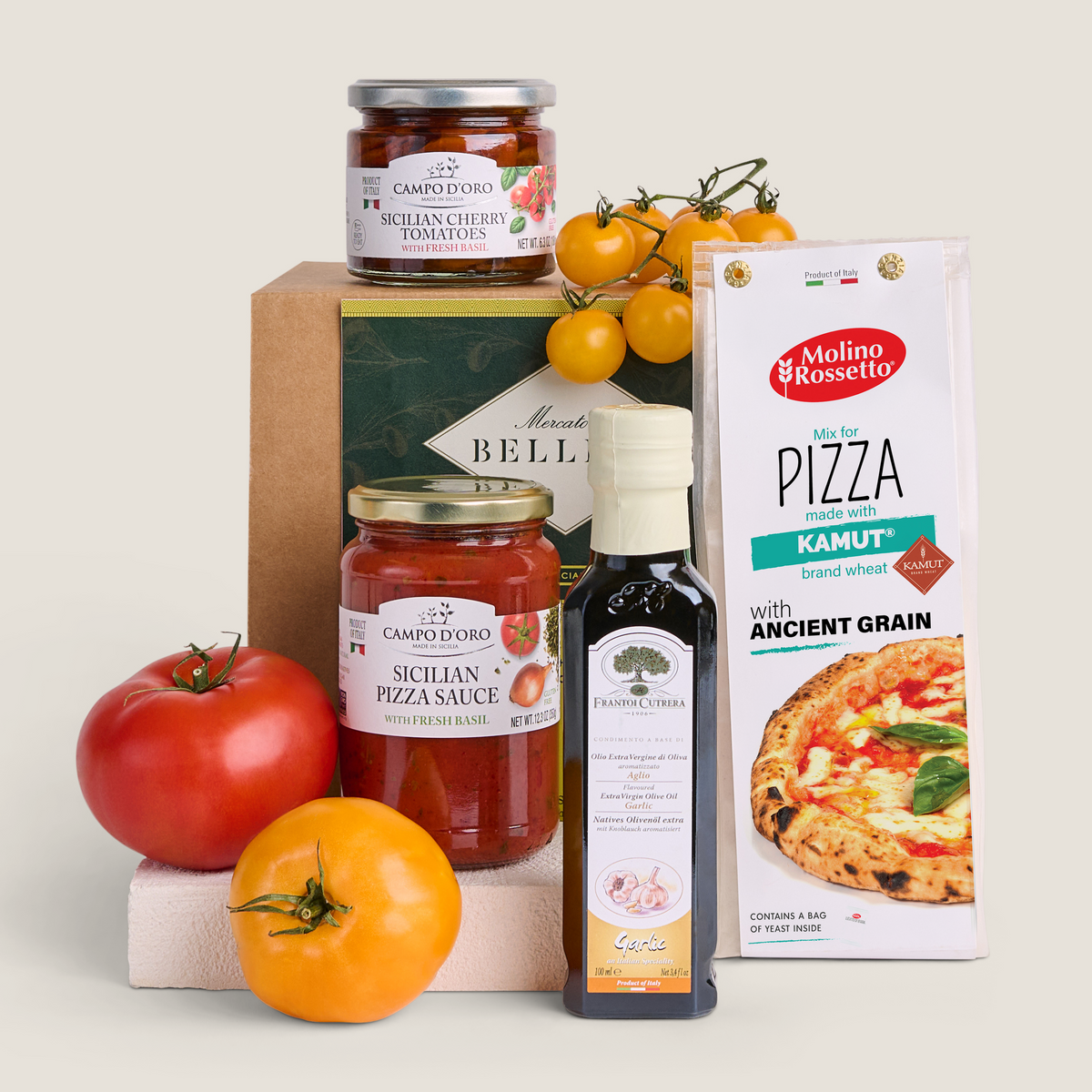 A styled product arrangement of food items on a light warm gray background: a jar of Campo d'Oro Sicilian Cherry Tomatoes, a bottle of Extra virgin Olive Oil, a box of Molino Rossetto Kamut Whole Wheat Pizza Flour, and fresh tomatoes and a vine of yellow tomatoes. No people or real environment are present, so this is Product/Packaging.