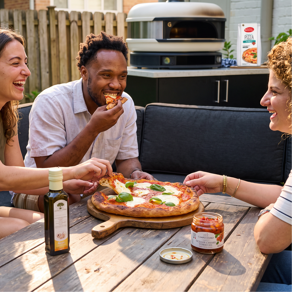 Three people laughing around a wooden table outdoors, sharing a freshly baked pizza with a jar of marinara sauce and a bottle of olive oil in the foreground. In the background, a person uses a white outdoor pizza oven on a black patio table.