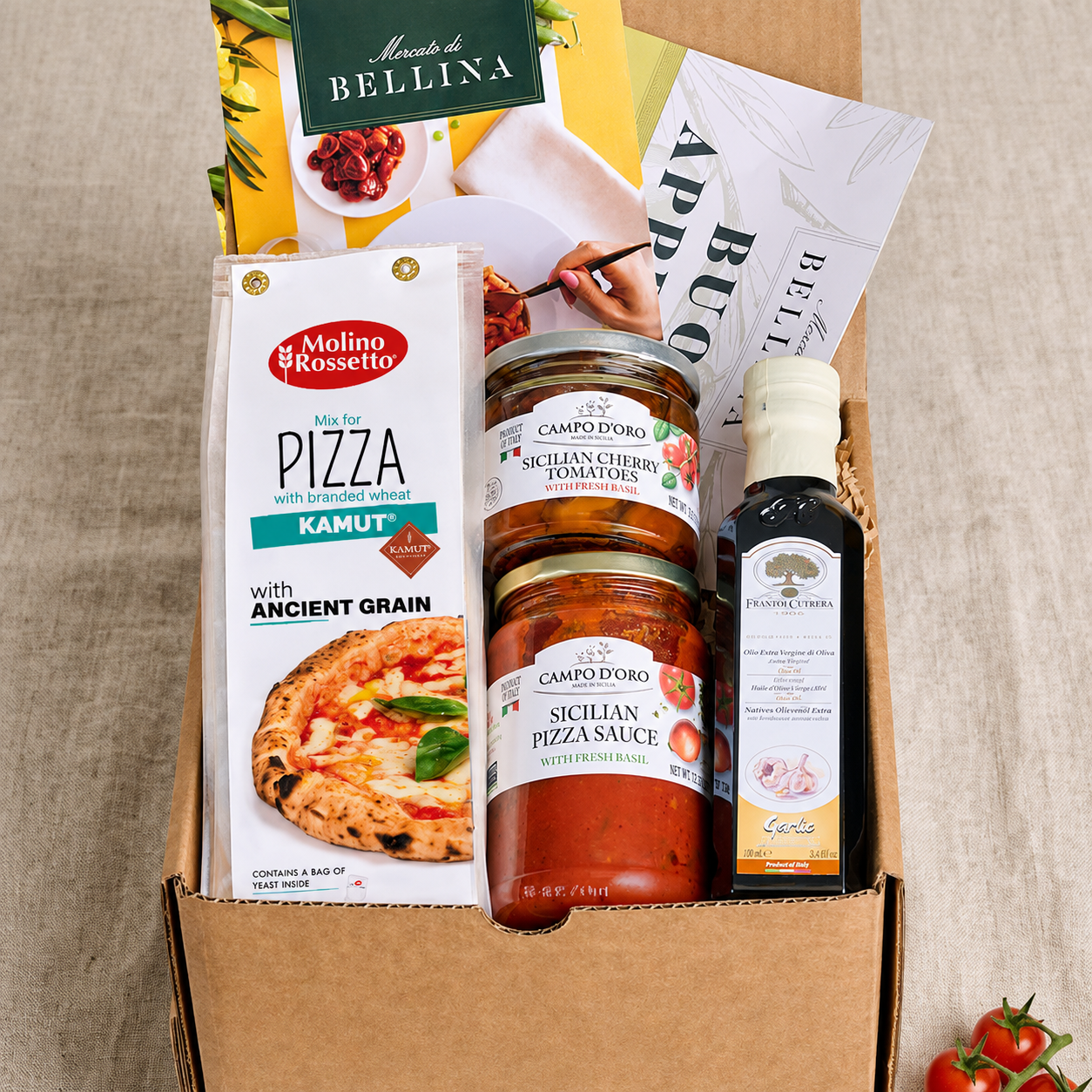 A curated pizza gift box filled with food products, including a package of Molino e Rossetto pizza, jars of Campano pizza sauce, a bottle of balsamic vinegar, and a booklet by Il Cibo di Bella. The box is styled on a beige linen background with fresh cherry tomatoes in the corner.