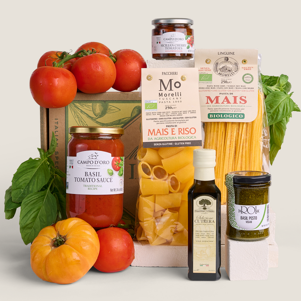A curated Italian food gift set arranged on a light beige background, including fresh tomatoes, basil leaves, a bottle of olive oil, a jar of basil pesto, a box of Mo Moretti pasta, a bag of Mais E Riso pasta, and a jar of pasta sauce.