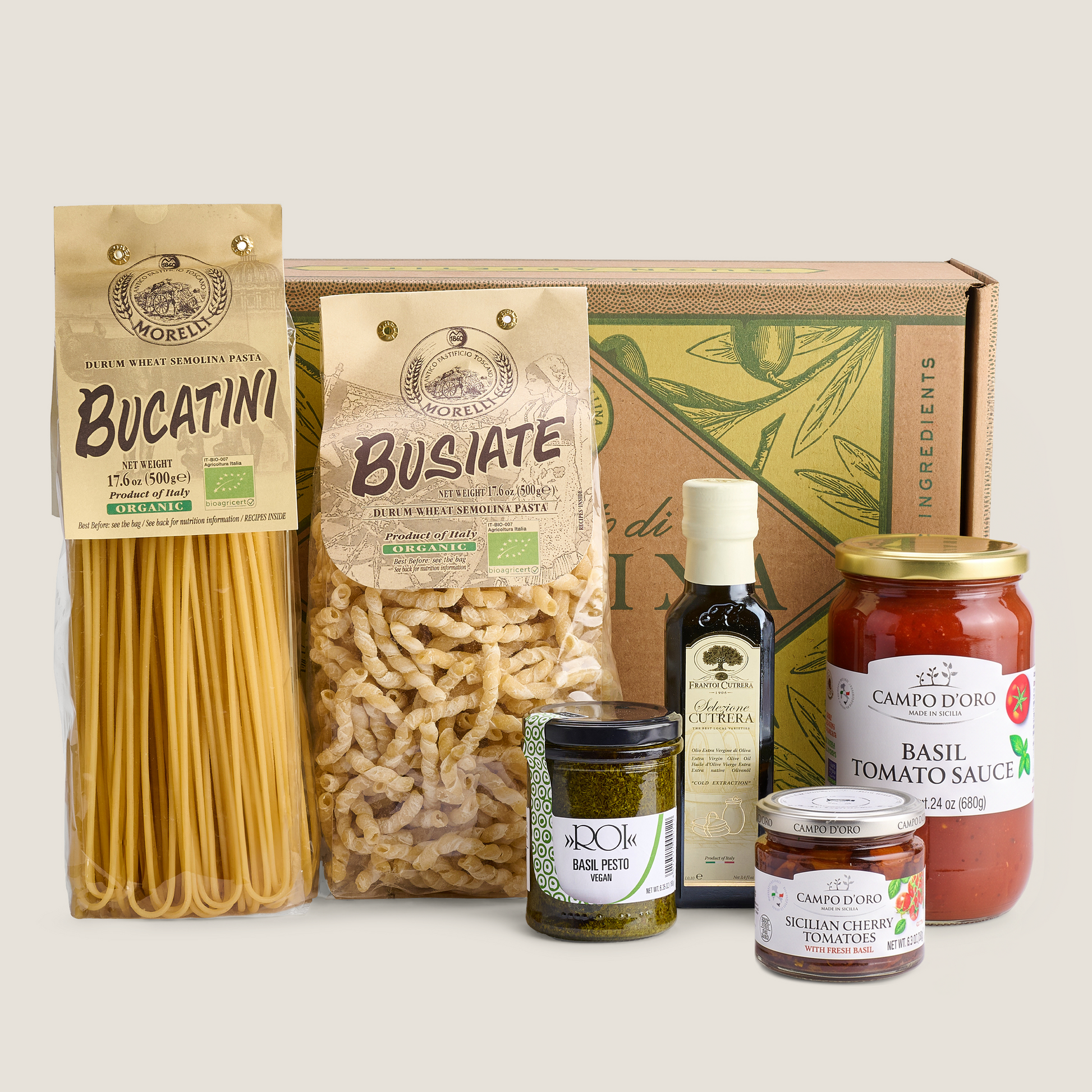 A curated food gift set featuring pasta, sauces, and spreads arranged in front of a branded cardboard box on a light beige background. Visible items include a bag of Bucatini pasta, a bag of Busate pasta, a bottle of Balsamic Vinegar, a jar of Basil Pork, a jar of Sicilian Cucumber Tomato, and a jar of Campoliano Basil Tomato Sauce.