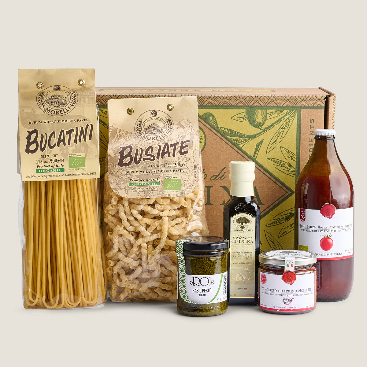 A curated food gift set featuring pasta, sauces, and spreads arranged in front of a branded cardboard box on a light beige background. Visible items include a bag of Bucatini pasta, a bag of Busiate pasta, a bottle of Balsamic Vinegar, a jar of Basil Pesto, a jar of Sicilian Cucumber Tomato, and a jar of  Basil Tomato Sauce.
