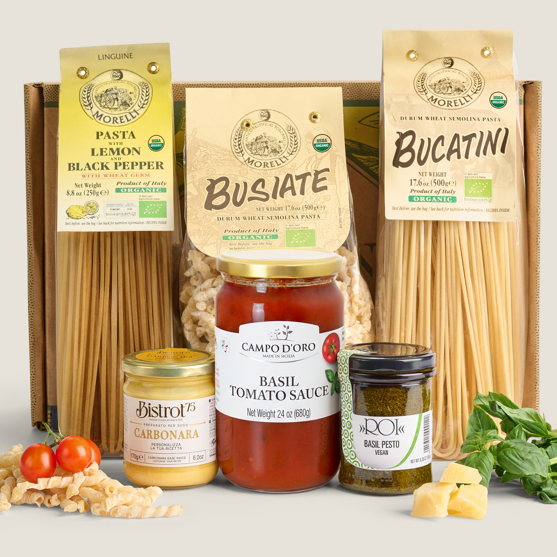 A curated pasta and sauce gift set arranged on a light neutral background. Three large boxes of pasta are visible behind the items, including labels for Pasta Lemon Black Pepper, Busiate, and Bucatini.