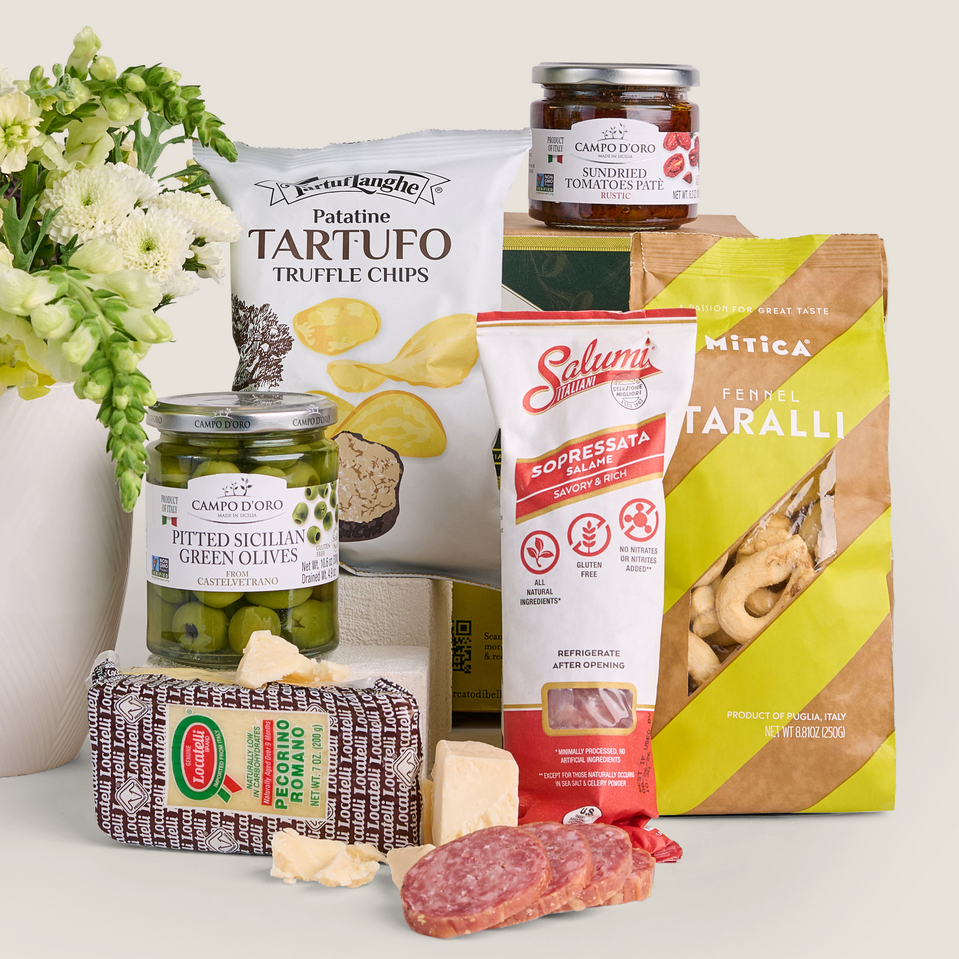 A curated assortment of Italian snacks and cured foods arranged on a white surface: a bag of Truffle chips, a jar of Campodoro Pitted Sicilian Green Olives, a jar of sundried tomatoes, a package of Salumi  Sopressata, a bag of Mita Ferrel Taralli, a block of pecorino romano, sliced salami, and a vase of white flowers in the background.