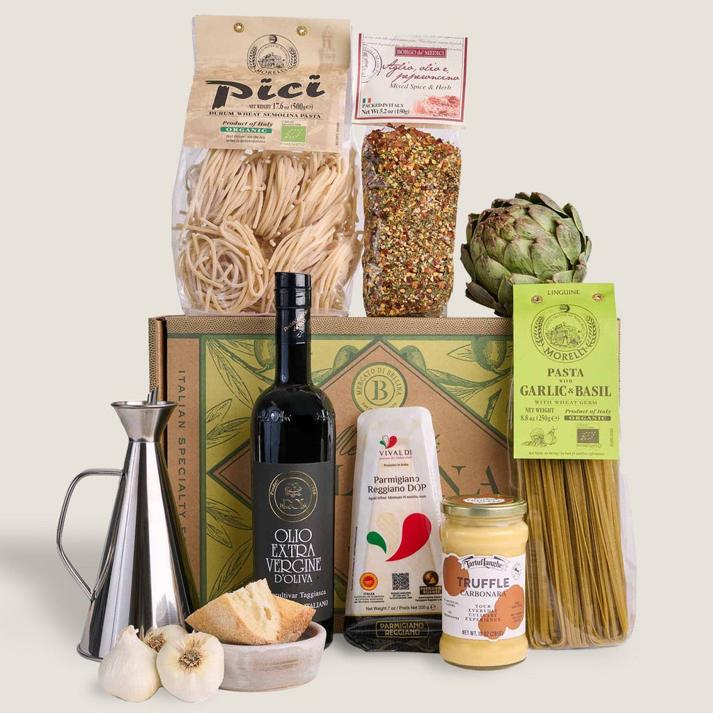 A curated gourmet food gift set arranged in front of a decorative box on a light beige background. The set includes Pici spaghetti, a bag of colorful pasta, an artichoke, a bottle of olive oil, a jar of ragù al pomodoro, a package of garlic and basil pasta, a metal measuring cup, garlic bulbs, and a wedge of cheese.