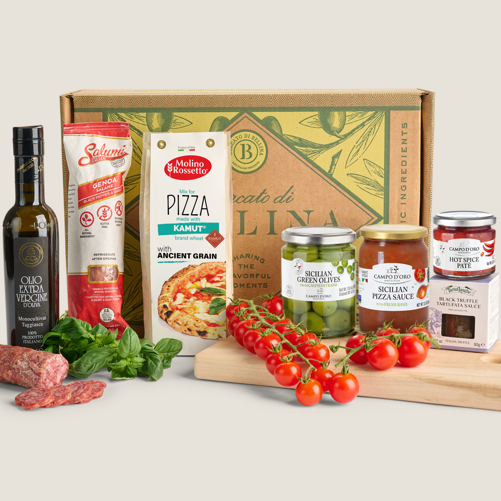 A curated food gift set arranged in front of a branded cardboard box on a light neutral background. Visible items include a bottle of olive oil, a tube of Salumi, a box of pizza dough, jars of pickled greens and pizza sauce, hot sauce, and boxes of cheeses, with sliced salami, basil, and cherry tomatoes styled around the products.