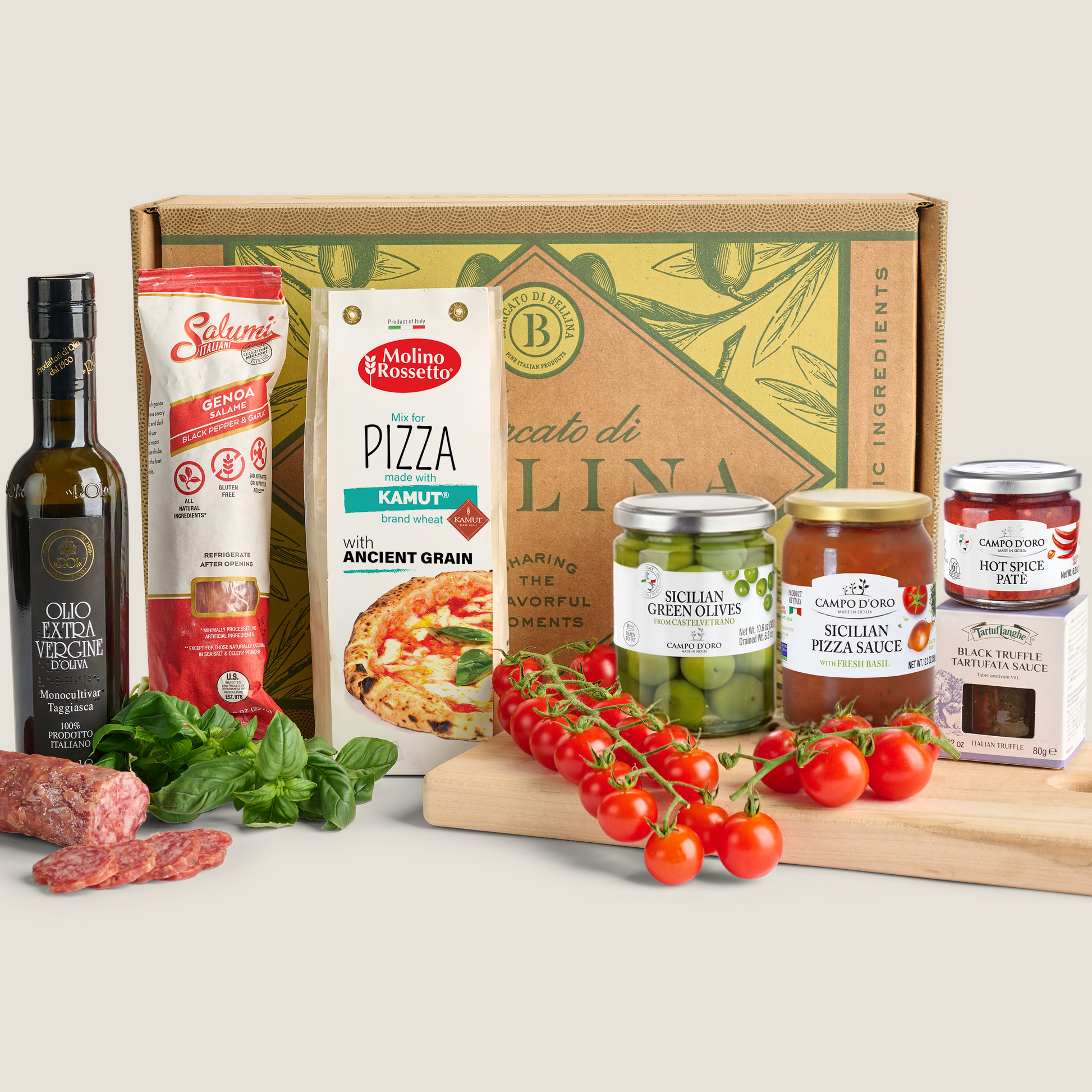 A curated food gift set arranged in front of a branded cardboard box on a light neutral background. Visible items include a bottle of olive oil, a tube of Salumi, a box of pizza dough, jars of pickled greens and pizza sauce, hot sauce, and boxes of cheeses, with sliced salami, basil, and cherry tomatoes styled around the products.