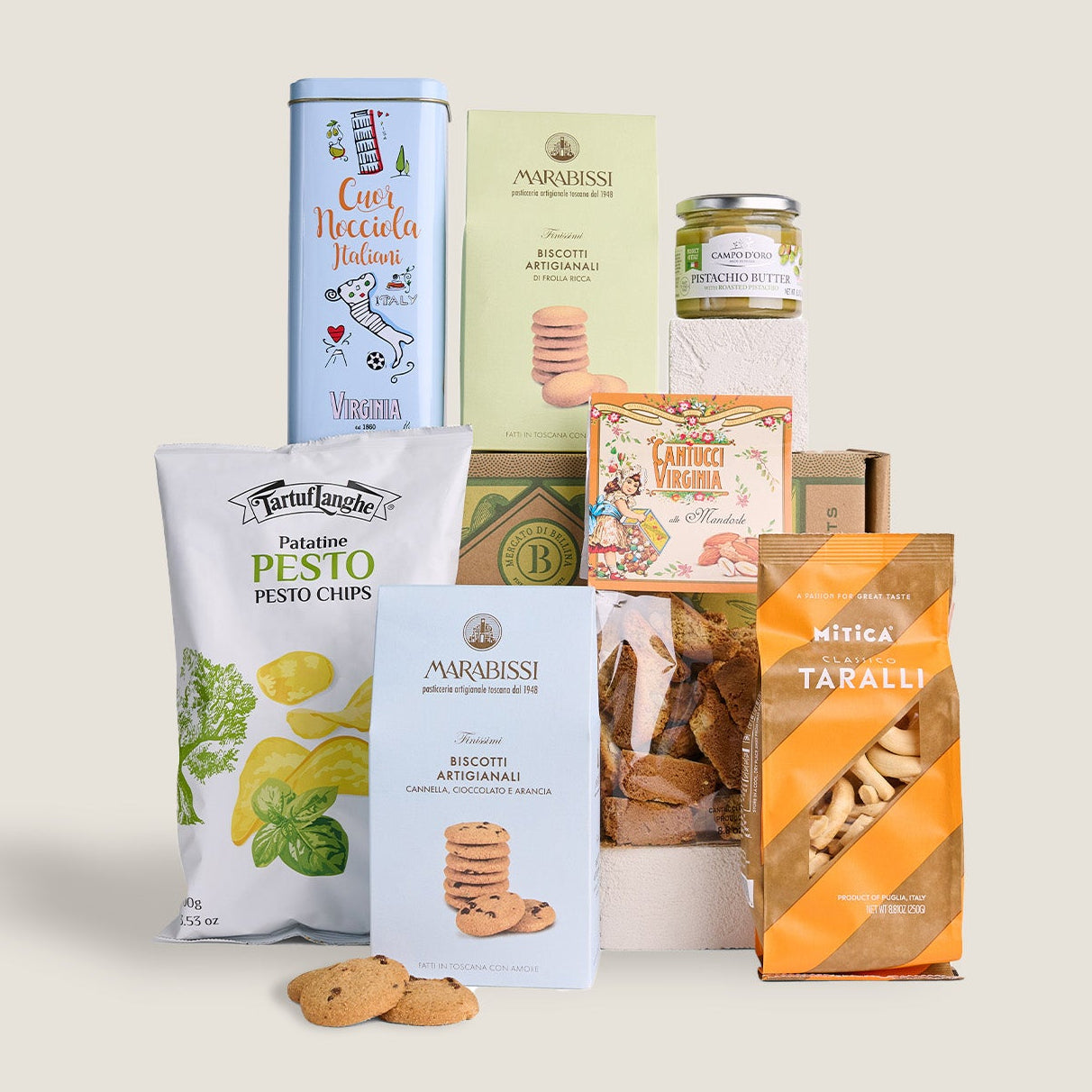 A curated gift basket of Italian snacks and sweets, featuring a tin of Cava Niccola Moscato, a bag of Pesto Chips, a box of Maraussi Biscotti, a jar of honey, and a bag of Taralli, arranged on a neutral beige background.