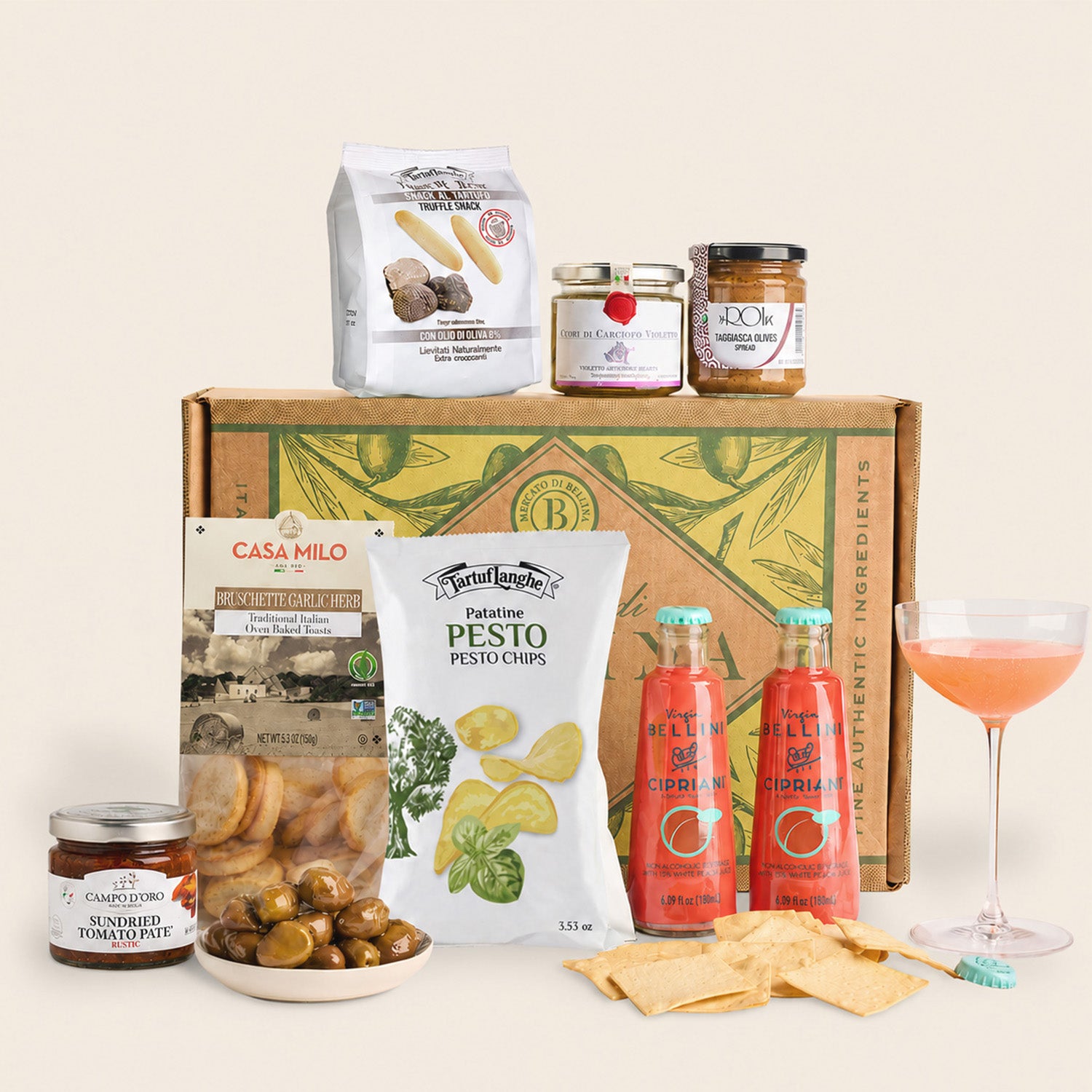 A curated gourmet gift basket featuring a brown cardboard box with green botanical illustrations, surrounded by various food items including a bag of pasta, pesto, crackers, olives, hot sauce, and a pink cocktail glass on a light beige background.