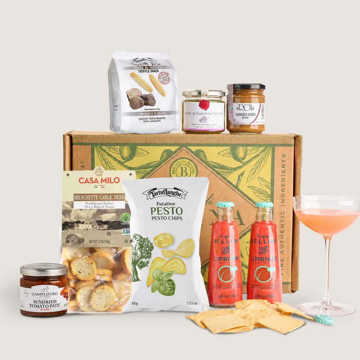 A curated snack and appetizer gift set arranged in front of a branded cardboard box on a light beige background. The set includes a bag of pasta, two jars of spreads, a bag of pasta with anchovies, a bag of potato chips with broccoli and pesto, two bottles of hot sauce, crackers, and a pink cocktail glass.