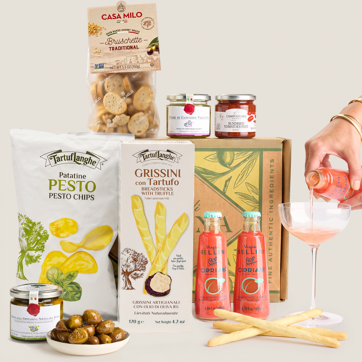 A curated food gift assortment on a light warm-gray background, including a bag of Tartuflanche Patatine Pesto Chips, a box of Grissini with truffle, jars from Casa Milo, two bottles of Bellini organic salami, a bowl of olives, breadsticks, and a hand pouring pink drink into a coupe glass.