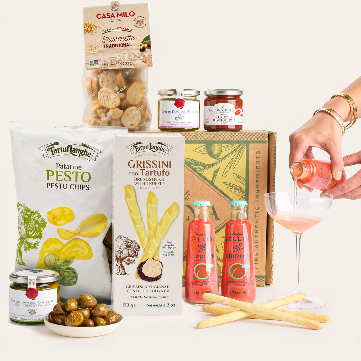 A curated food gift assortment on a light warm-gray background, including a bag of Tartuflanche Patatine Pesto Chips, a box of Grissini with truffle, jars from Casa Milo, two bottles of Bellini organic salami, a bowl of olives, breadsticks, and a hand pouring pink drink into a coupe glass.