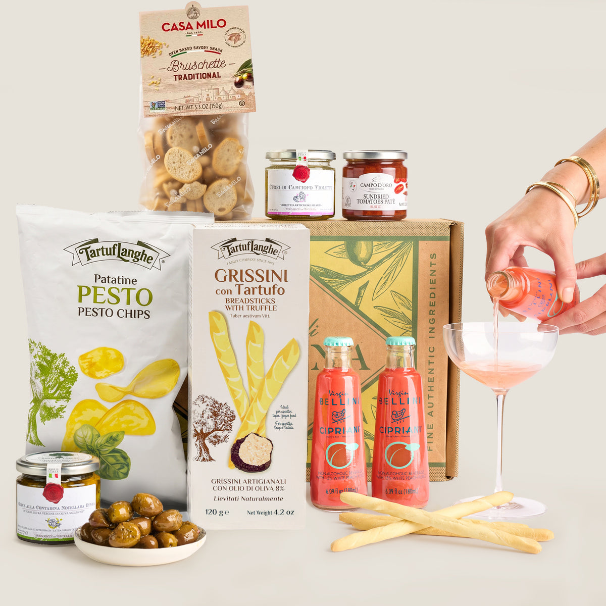 A curated food gift assortment on a light warm-gray background, including a bag of Tartuflanche Patatine Pesto Chips, a box of Grissini with truffle, jars from Casa Milo, two bottles of Bellini organic salami, a bowl of olives, breadsticks, and a hand pouring pink drink into a coupe glass.