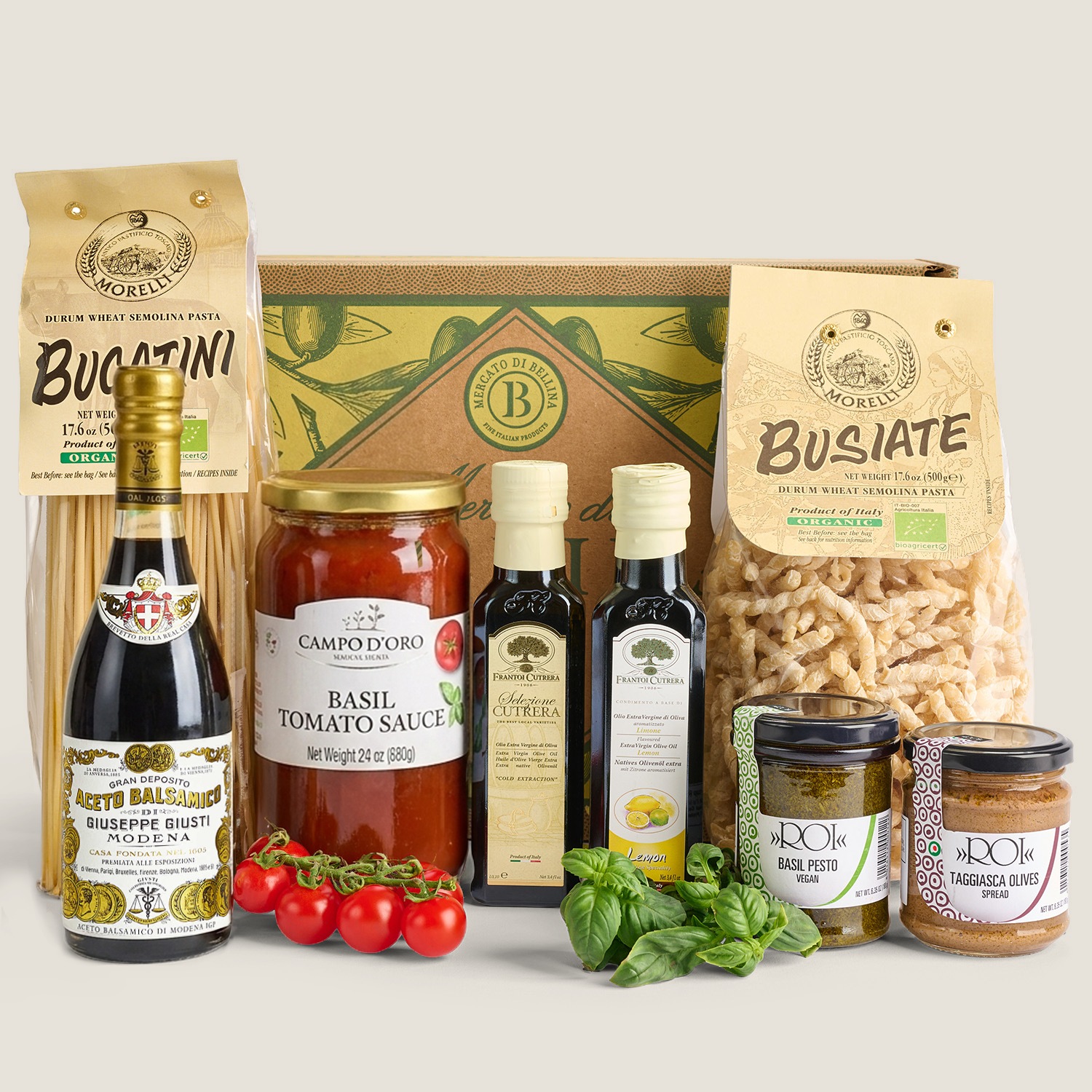 A curated gourmet food gift set arranged on a light beige background. The set includes a box of Bucatini semolina pasta, a bag of busiate pasta, a bottle of BuGATINI ragù alle vitello muscoli spaghetti, a bottle of Campo d'Oro Italiano Basil Tomato Sauce, bottles of olive oil and vinegar, jars of spreads, and fresh basil and cherry tomatoes.