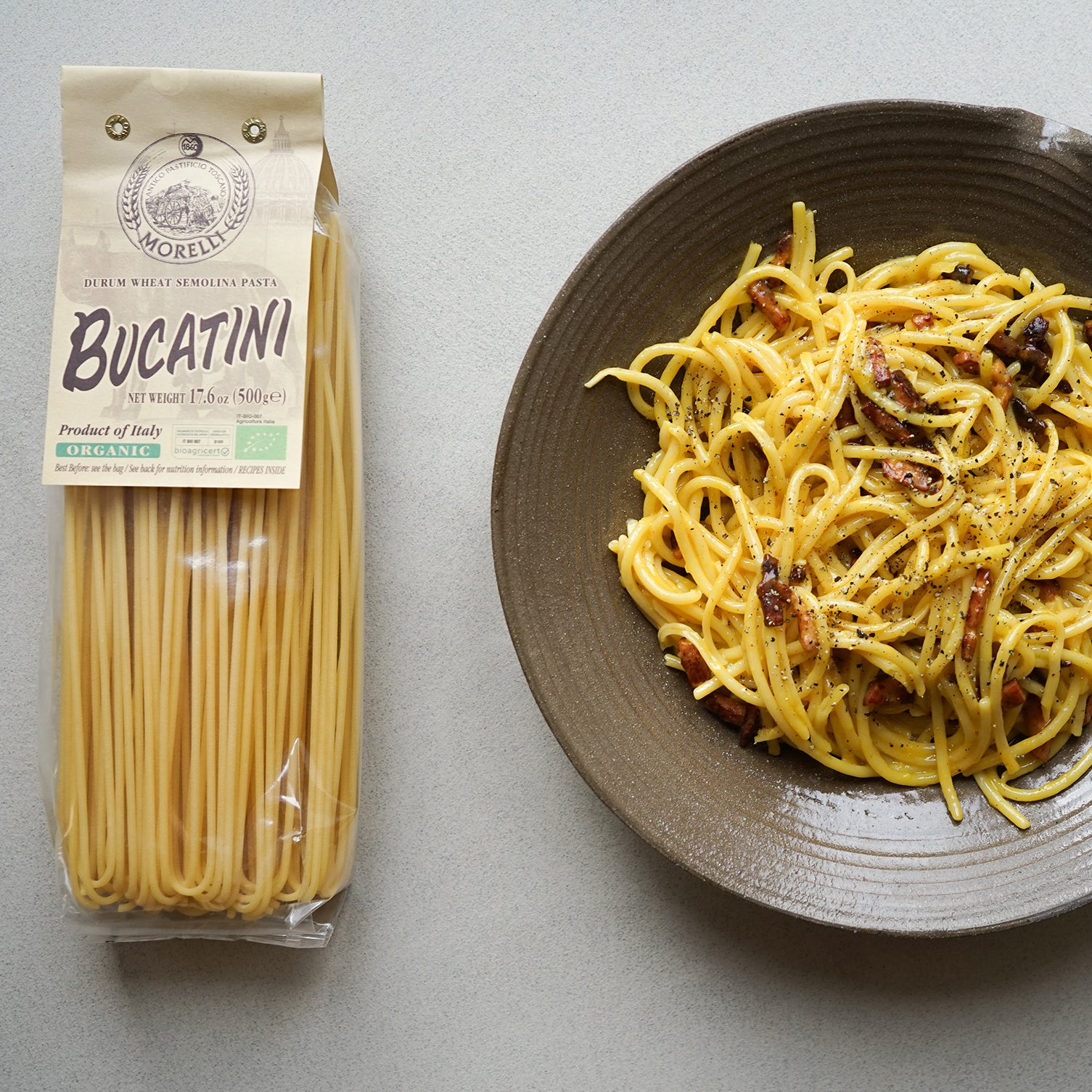Organic bucatini pasta package next to a plate of carbonara with pancetta