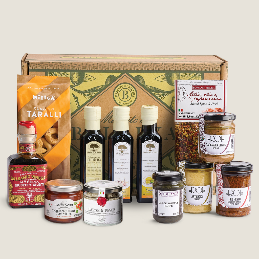 A curated gourmet gift box set featuring olive oil, balsamic vinegar, spreads, and snacks arranged in front of a branded gift box on a light beige background.
