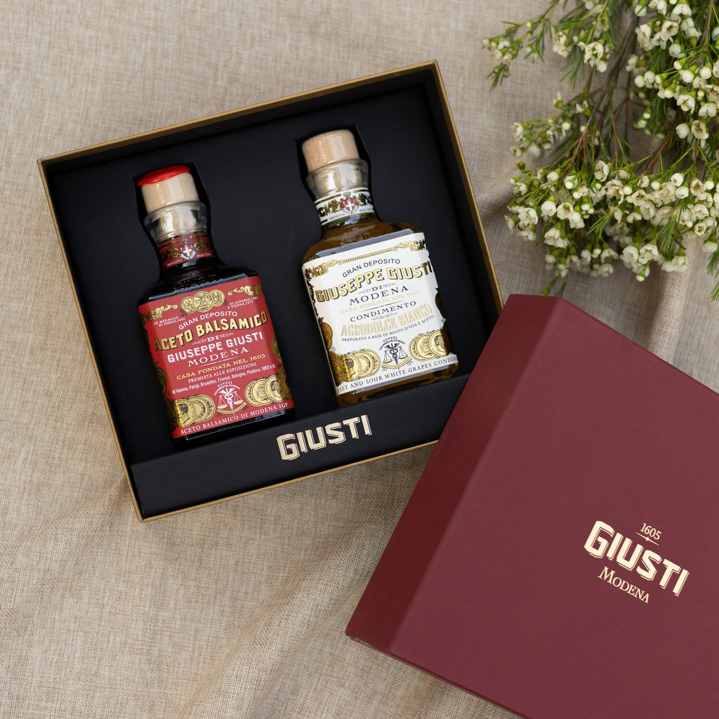 Giusti & Cibo gift set featuring two small bottles of balsamic and white wine vinegar in a black presentation box, with the burgundy Giusti & Cibo box lid placed beside it on a beige linen fabric background.
